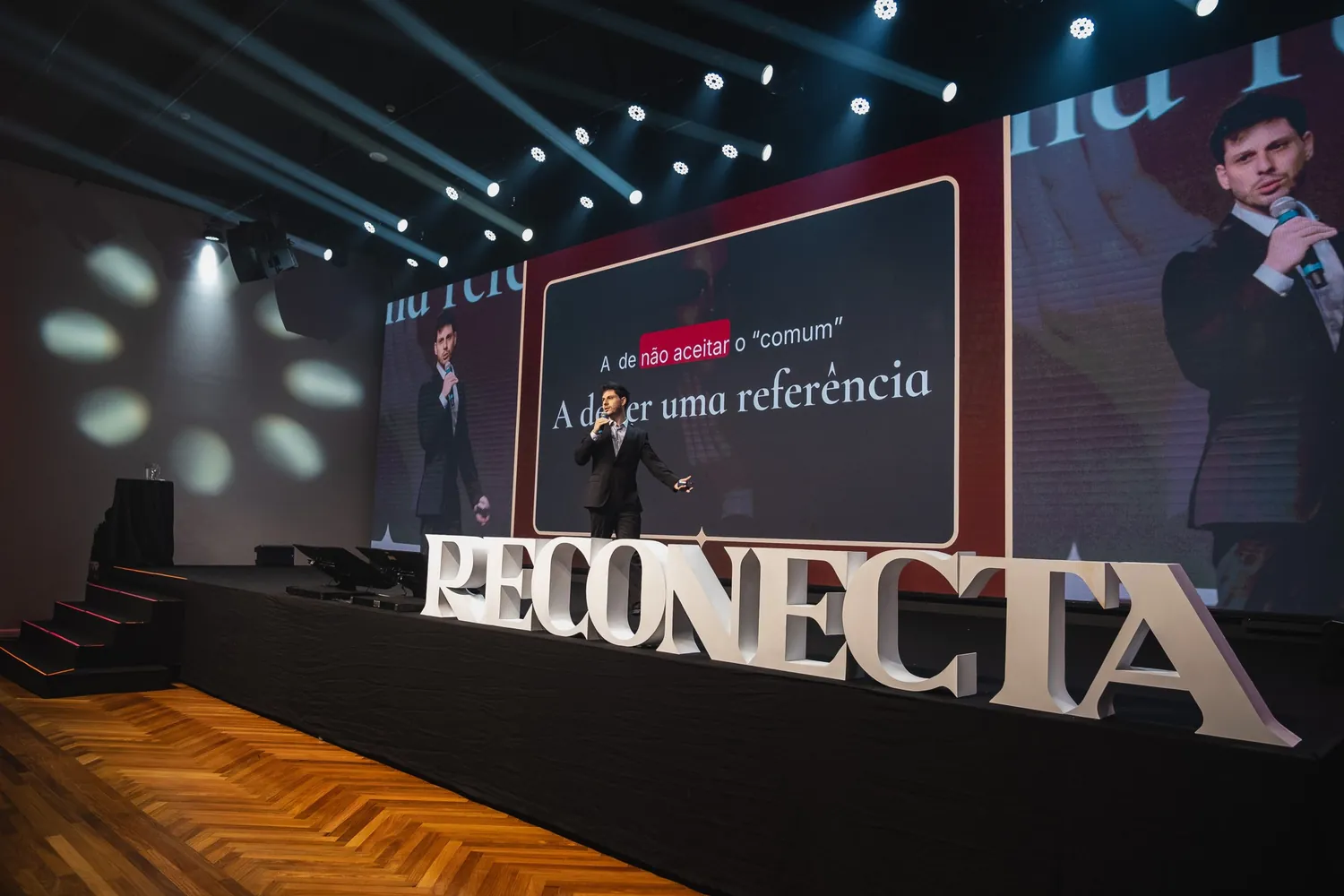 Palco do evento presencial da Reconecta: condutor diante do telão gigante, com o letreiro RECONECTA na boca do palco